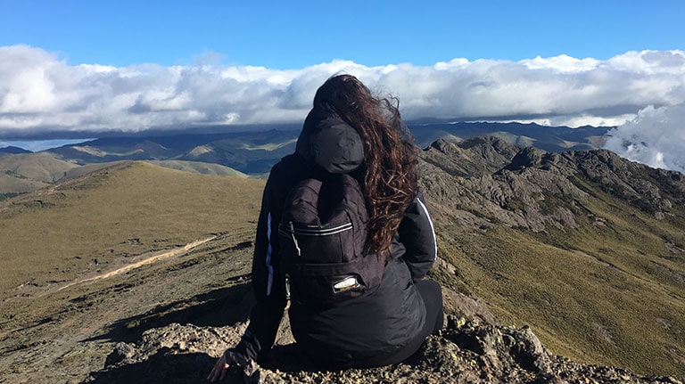A person wearing a backpack stands on a rocky hilltop and looks over a landscape of wild, grassy valleys.